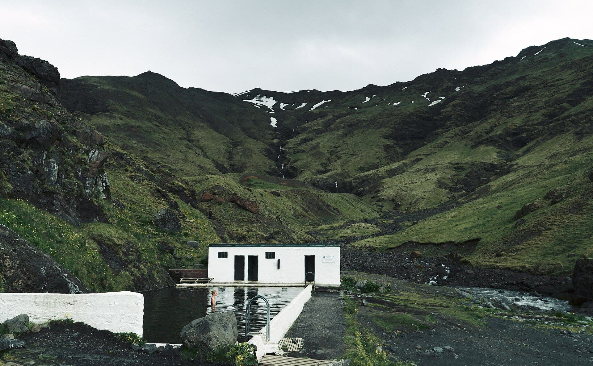 A small white building beside an outdoor pool in a green mountain valley.